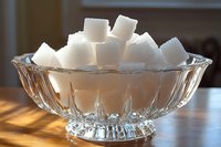 White sugar cubes stacked in elegant crystal glass bowl on wooden table with natural sunlight creating dramatic shadows and reflections through faceted glass.