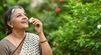 Senior woman with gray hair and South Asian ethnicity enjoying fresh flower in blooming garden, expressing joy and wellness outdoors.