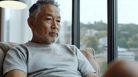 Senior Asian man with gray beard wearing casual gray t-shirt sitting thoughtfully near window at home, looking away with contemplative expression. Natural daylight setting.