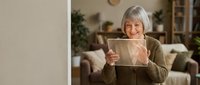 Happy senior Caucasian woman with gray bob haircut holding digital tablet in cozy living room, smiling while using technology at home for communication.