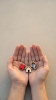 Hands holding wooden house, medical cross, and shield symbols on neutral background, representing home insurance, healthcare coverage, and family protection concepts.