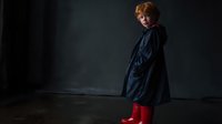 Young redhead child in dark raincoat and bright red boots standing against moody dark background, looking over shoulder with curious expression.