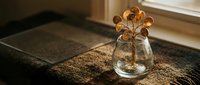 Small glass vase with dried golden flowers on woven placemat by window creating warm natural home decor atmosphere for interior design projects.