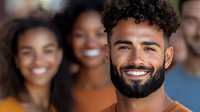 Young mixed-race man with curly hair and beard smiling confidently at camera with diverse group of happy people in background for teamwork concepts.