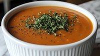 Creamy tomato soup garnished with fresh chopped herbs and black pepper in white ceramic bowl, close-up view showing smooth texture and vibrant orange color.