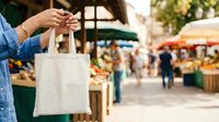 Close-up of person in denim shirt holding blank white canvas tote bag at farmers market with produce stalls and shoppers in background.