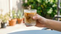 Hand holding glass of iced cola with ice cubes outdoors in sunny garden setting, refreshing cold beverage for summer drinks and lifestyle photography.