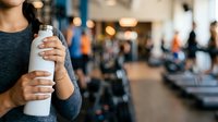 Woman holding white water bottle at gym with blurred fitness equipment and people in background, suitable for health, wellness, hydration and sports lifestyle marketing.