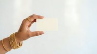 Hand holding blank cream colored card against white background, wearing gold bracelets. Mockup for business cards, invitations, or promotional materials.