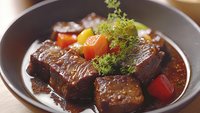 Tender braised beef chunks with cherry tomatoes and fresh herbs in dark savory sauce, served in black ceramic bowl on wooden surface. Close up food photography.