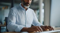 Mature businessman with gray beard working on computer keyboard in modern office environment, focused on digital task.