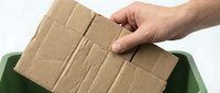 Caucasian male hand placing a flattened corrugated cardboard piece into a green recycling bin, promoting eco-friendly waste management.