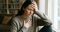 Young Asian woman experiencing stress and anxiety sitting by window in cozy home interior with hand on forehead showing mental health struggles and emotional distress.
