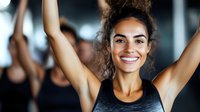 Young mixed race woman with curly hair smiling during workout at gym, wearing black tank top, arms raised up, expressing joy and achievement in fitness.