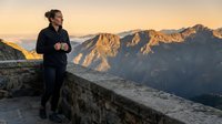 Middle-aged Caucasian male athlete jogging on mountain ridge at sunrise, wearing black athletic gear, with alpine peaks and golden light in background. Fitness and outdoor training concept.