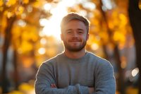 Young Caucasian man with beard wearing grey sweater smiling confidently with arms crossed against blurred autumn background with golden bokeh lights and foliage.
