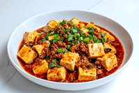 Spicy mapo tofu with minced meat and green onions in white bowl on marble surface, traditional Chinese Sichuan cuisine with red chili sauce and soft tofu cubes.
