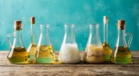 Various glass bottles and containers with different oils and liquids arranged on wooden surface against turquoise background for cooking and culinary use.
