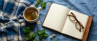 Cozy flat lay with herbal tea, open notebook, reading glasses, and mint leaves on a soft blue linen background for lifestyle and wellness content.