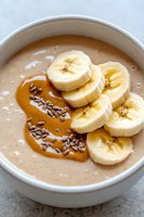 Creamy smoothie bowl topped with fresh banana slices, caramel sauce and chocolate sprinkles in white ceramic bowl on marble counter, healthy breakfast.