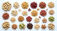 Assorted legumes and nuts arranged in circular piles on white background, including peanuts, cashews, chickpeas, red beans, pinto beans, and pistachios for food, nutrition, and cooking projects.