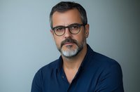 Middle-aged Caucasian man with gray beard and glasses wearing navy blue polo shirt against soft gray background, professional headshot style portrait.