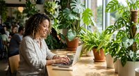 Young African American woman working on laptop in bright plant-filled home office space surrounded by green houseplants creating productive workspace atmosphere.