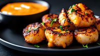 Pan-seared scallops with golden caramelized crust served on dark plate with orange sauce and fresh herbs for gourmet seafood dining experience.
