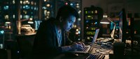 Young Asian male programmer coding on laptop in dark office with city lights background at night, focused developer working late hours on software project.