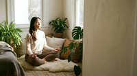 Young Asian woman practicing meditation in cozy living room with houseplants creating peaceful mindful atmosphere for wellness and relaxation.