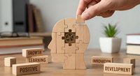 Hand placing final puzzle piece into wooden head silhouette surrounded by motivational blocks labeled growth, learning, positive, innovation, development.