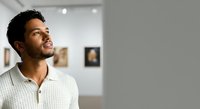 Man with dark curly hair and medium brown skin tone wearing white ribbed shirt, looking upward thoughtfully in modern gallery or office space with blurred background.