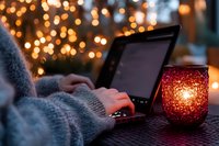 Person typing on laptop outdoors at dusk with glowing red candle holder and festive bokeh lights creating cozy autumn or winter atmosphere.