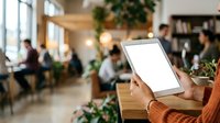 Person in orange sweater holding tablet with blank screen in modern office workspace with colleagues and plants.