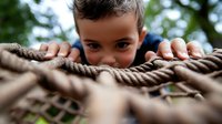 Young boy peeking over thick brown rope on outdoor playground equipment, playful expression with curious eyes visible above climbing structure.