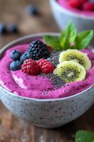 Pink smoothie bowl topped with fresh berries, kiwi slices, chia seeds and mint leaves in ceramic bowl. Healthy breakfast or snack with vibrant colors and textures.