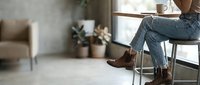 Person sitting at a window table in ripped jeans and brown leather ankle boots, relaxing in a cozy modern cafe with plants and a coffee cup.