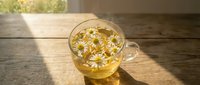 Glass mug of chamomile tea with floating daisy flowers on wooden table in warm sunlight for relaxation and wellness concepts.