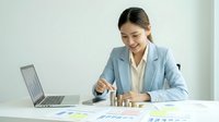 Young Asian businesswoman in light blue suit analyzing financial data and charts at modern office desk with laptop for business planning and strategy.