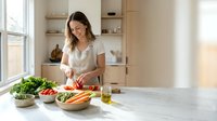 Young woman of mixed ethnicity wearing glasses and a white short-sleeved shirt, chopping fresh vegetables in a bright, minimalist kitchen. Healthy cooking and meal preparation.