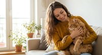 Young woman with long brown hair wearing mustard yellow sweater cuddling orange tabby cat on cozy living room sofa by sunny window with houseplants.