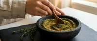 Person grinding fresh herbs with wooden spoon in stone mortar and pestle on kitchen counter for natural cooking and food preparation.