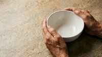 Senior adult with dark skin tone holding empty white ceramic bowl on textured beige surface, depicting poverty, hunger, need, charity, or food insecurity concept for humanitarian campaigns.
