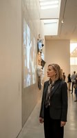 Middle-aged woman with shoulder-length blonde hair wearing dark blazer and gray scarf viewing digital art installation on gallery wall in contemporary museum space with skylights.