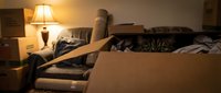 Curious tabby cat hiding inside an open cardboard box surrounded by moving boxes in a dimly lit living room, peeking with bright green eyes.