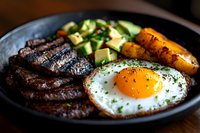 Grilled steak and sunny side up egg served with fresh avocado and roasted carrots on black plate, close up view of protein-rich breakfast or brunch meal.