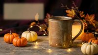 Rustic ceramic mug with warm beverage surrounded by decorative mini pumpkins and autumn branches with glowing string lights for fall seasonal decor and holiday entertaining.