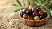 Fresh mixed olives in wooden bowl on burlap with olive leaves, natural light setting. Mediterranean cuisine ingredients for gourmet food styling.