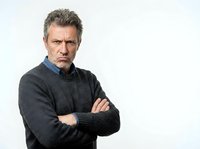Mature man with gray hair and facial stubble wearing dark gray sweater over blue shirt, arms crossed, serious expression, isolated on white background for corporate and professional use.