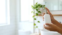 Woman with medium skin tone holding white cosmetic pump bottle with gold accents in bright modern interior with potted plant, suitable for beauty, skincare, and wellness marketing.